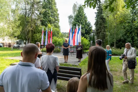 Uroczystość z flagami, przemawiająca kobieta i zgromadzenie osób w parku.