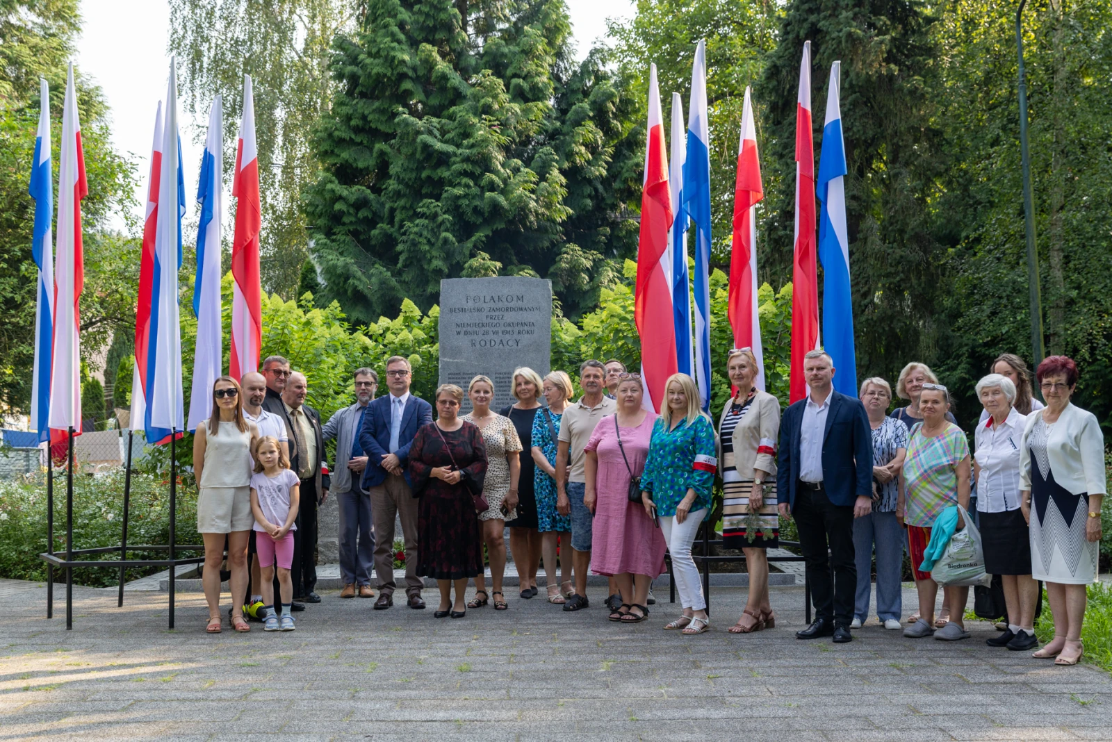 Grupa ludzi w strojach formalnych i codziennych, stojąca przed pomnikiem otoczonym flagami.  Autor: Bogusław Świerzowski