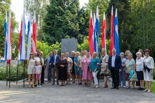 Grupa ludzi w strojach formalnych i codziennych, stojąca przed pomnikiem otoczonym flagami.