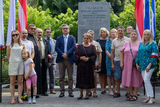 Grupa ludzi stojąca przy pomniku, z flagami w tle, podczas uroczystości upamiętniającej.