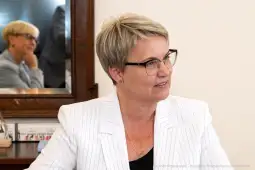 Kobieta w okularach, w białym żakiecie, rozmawia w biurze. W tle odbicie innej osoby.