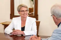 Kobieta w okularach, w białym garniturze, uśmiecha się podczas rozmowy przy stole.