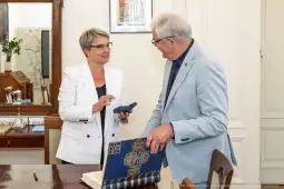 Kobieta w białym żakiecie rozmawia z mężczyzną w szarym garniturze przy stole.