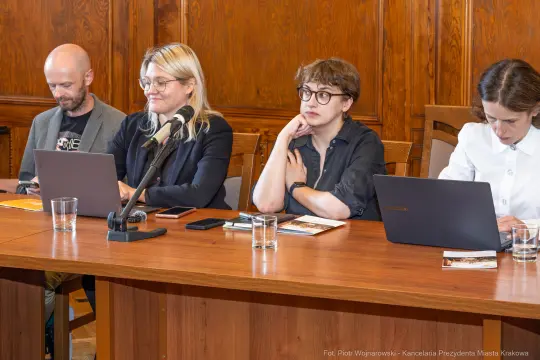 Cztery osoby siedzą przy stole, korzystając z laptopów i mikrofonu.