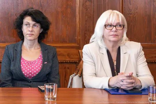 Dwie kobiety siedzące przy stole, w tle drewniana ściana.