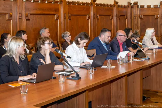 Spotkanie w sali obrad z uczestnikami przy stole, każdy z laptopem i mikrofonem.