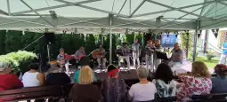 Zespół muzyczny grający na żywo pod zadaszeniem, publiczność siedząca na ławkach.