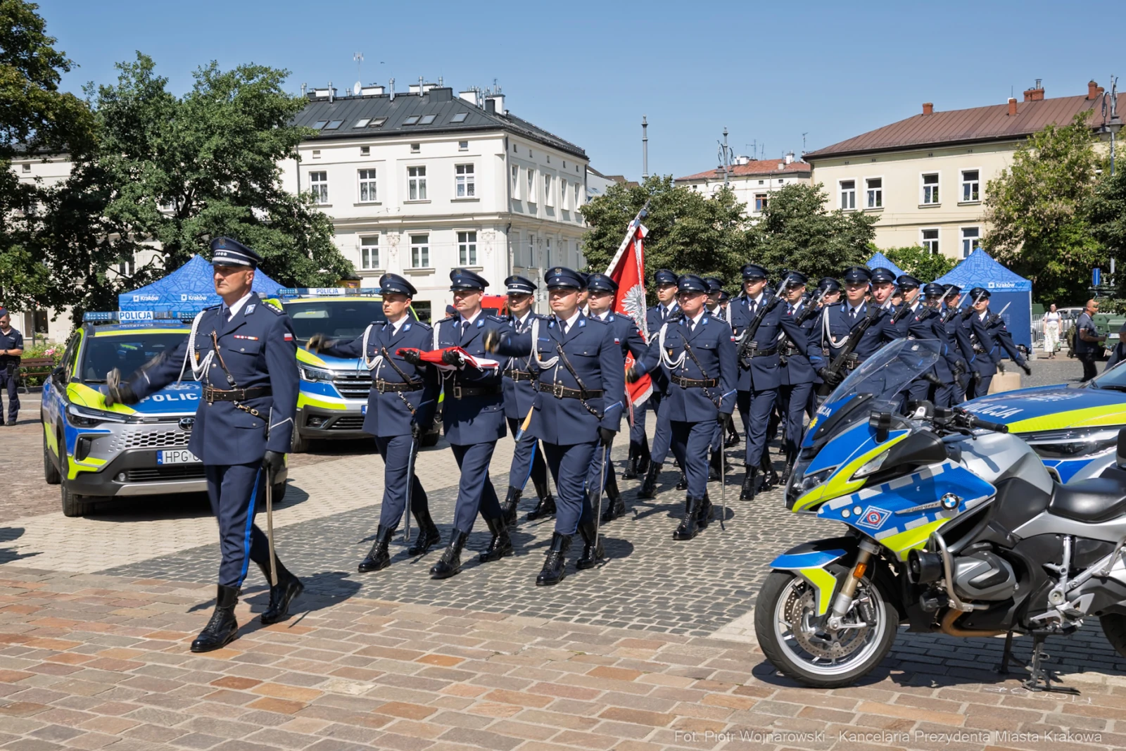 Parada policji z flagą, radiowóz i motocykl w tle, słoneczna pogoda.  Autor: Piotr Wojnarowski