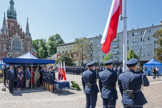 Uroczystość z udziałem mundurowych, flagi Polski i budynku w tle.