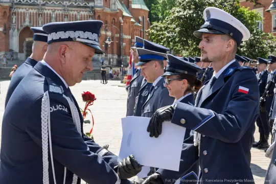 Ceremonia wręczenia dokumentów przez policjantów w mundurach na tle zabytkowej architektury.
