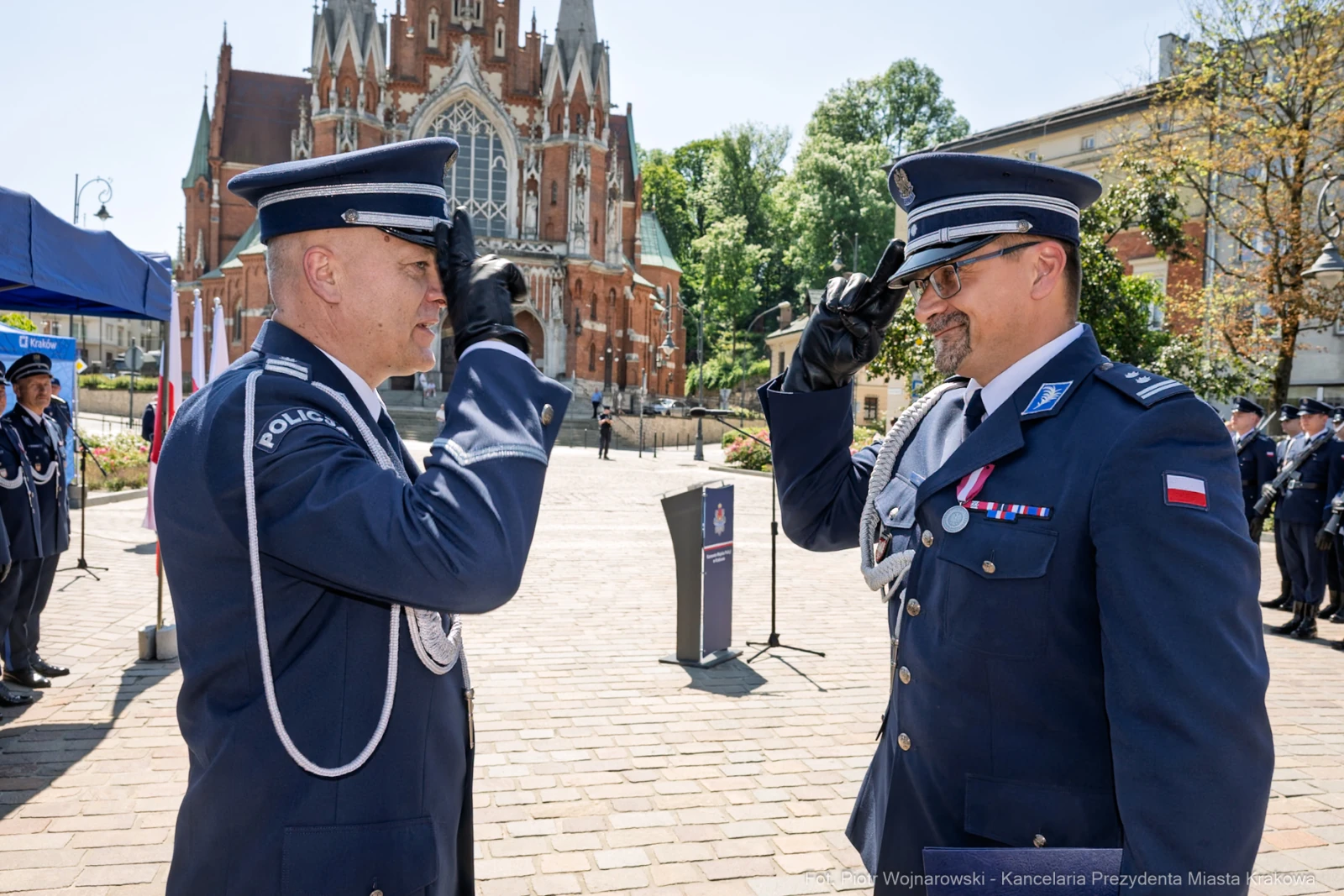 Dwaj policjanci w mundurach składają sobie salut przed budynkiem z czerwonej cegły.  Autor: Piotr Wojnarowski
