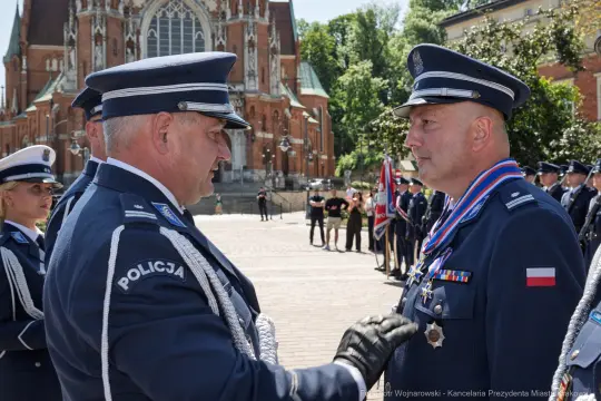 Dwaj policjanci w mundurach, jeden z odznaczeniami, rozmawiają na tle katedry.