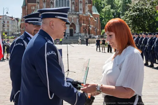 Wręczenie nagrody przez policjanta kobiecie na tle budynku i grupy policjantów.