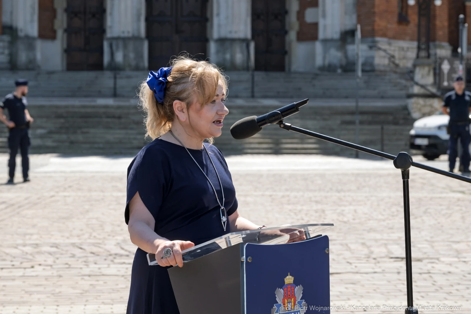 Kobieta przemawia do mikrofonu na tle budynku, w tle stoją policjanci.  Autor: Piotr Wojnarowski