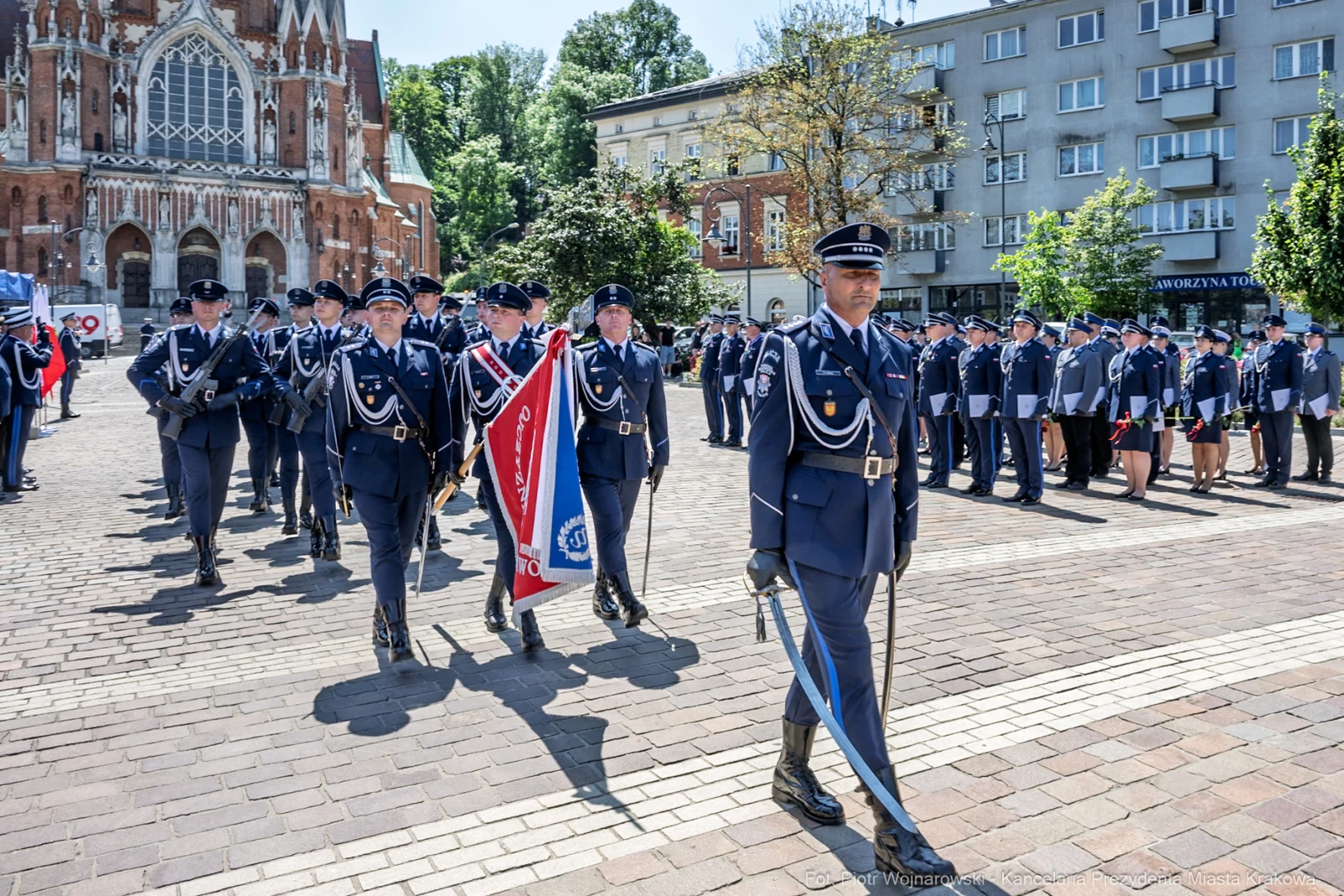 Parada wojskowa z uczestnikami w mundurach i sztandarem na tle budynków.  Autor: Piotr Wojnarowski