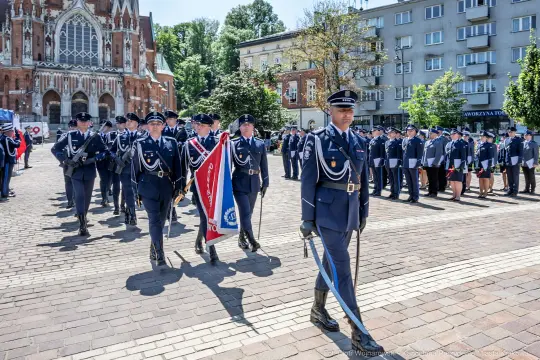 Parada wojskowa z uczestnikami w mundurach i sztandarem na tle budynków.