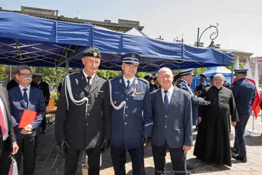 Trzech mężczyzn w mundurach oraz eleganckich strojach, stojących na tle namiotów i flag.
