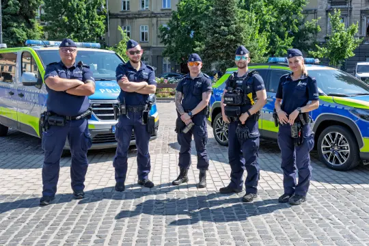 Grupa policjantów w mundurach stoi obok radiowozów na placu.