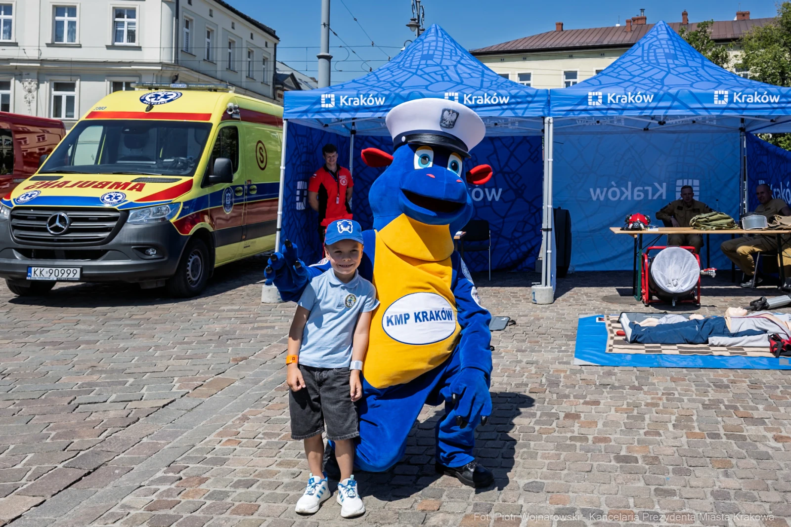 Dziecko pozujące z niebieską maskotką w kapeluszu na tle namiotów i karetki.  Autor: Piotr Wojnarowski
