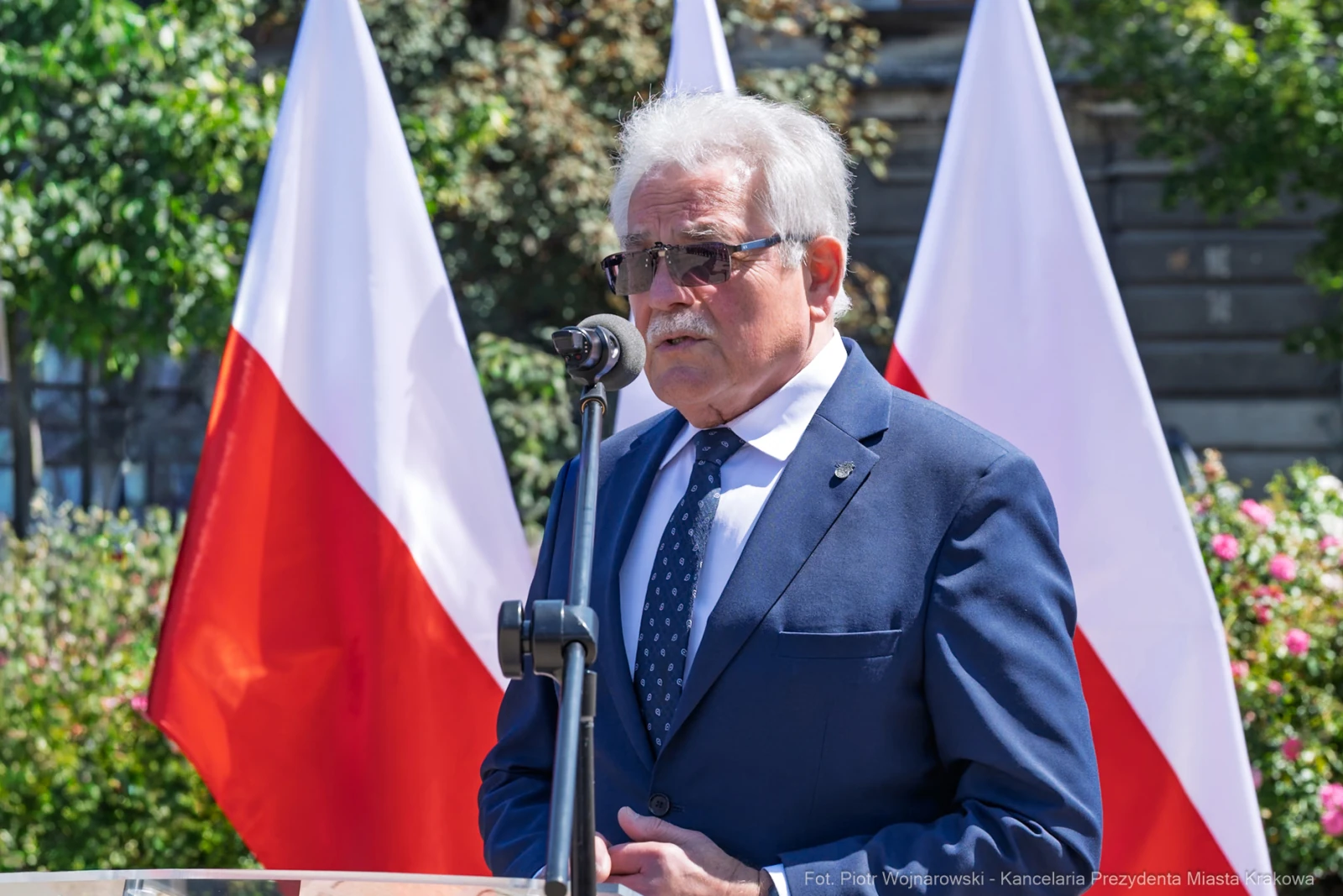Mężczyzna w okularach przeciwsłonecznych przemawia przy mikrofonie, w tle flagi Polski.  Autor: Piotr Wojnarowski