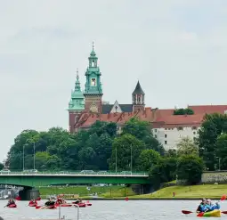 Zamek na Wawelu z wieżami i zielonymi kopułami, w tle rzeka i kajakarze.