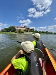 Dzieci w kajaku płyną w stronę zamku nad rzeką. Słoneczne niebo z chmurami.