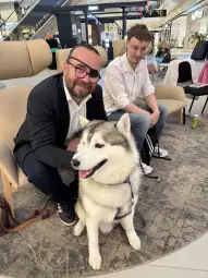 Mężczyzna w okularach z psem rasy husky, obok siedzi drugi mężczyzna.