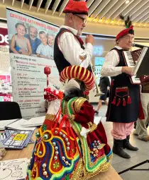 Dwaj mężczyźni w tradycyjnych strojach grają na instrumentach, obok kolorowa kukła.