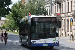 Elektryczny autobus na ulicy, obok rowerzyści i budynki w tle.