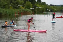 Kobieta na desce SUP, mężczyzna i dziecko na kajaku, tor do slalomu wodnego w tle.