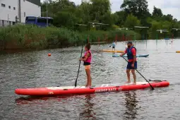 Dwoje ludzi na desce paddleboardingowej na wodzie, otoczeni zielenią i innymi kajakarzami.