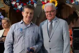 Dwaj mężczyźni w formalnych strojach, jeden z medalem, stoją obok siebie w dekoracyjnym otoczeniu.