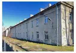 Budynek mieszkalny z szarym tynkiem i oknami, otoczony trawnikiem i płotem.