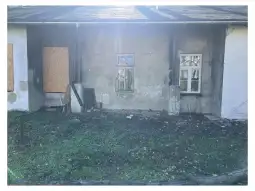 Zniszczona ściana budynku z zasłoniętymi oknami i zarośniętym terenem.