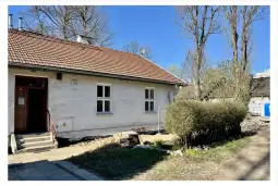 Biały dom z czerwonym dachem, obok krzewy i ścieżka, w tle niebo bez chmur.