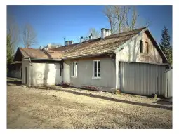 Stary dom z szarymi ścianami i dachówką, otoczony drzewami i pustym terenem.