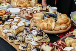 Stół z różnorodnymi ciastami i wypiekami, w tym babkami i bułkami.