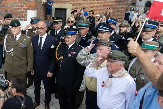 Grupa mężczyzn w mundurach i cywilnych strojach, salutujących podczas uroczystości.