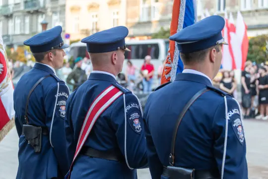 Trzech policjantów w mundurach, stojących z flagami na tle zgromadzenia ludzi.