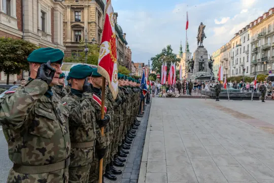 Żołnierze w mundurach salutują podczas uroczystości na placu z pomnikiem.