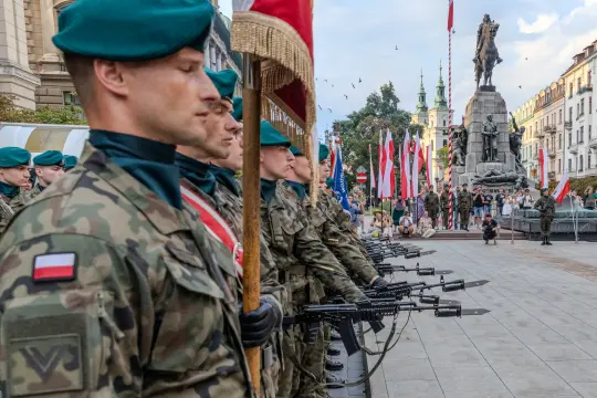 Żołnierze w mundurach wojskowych z bronią, stojący w szeregu podczas uroczystości z flagami.