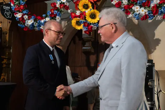 Mężczyźni w garniturach uścisną ręce podczas uroczystości, w tle kwiatowe dekoracje.