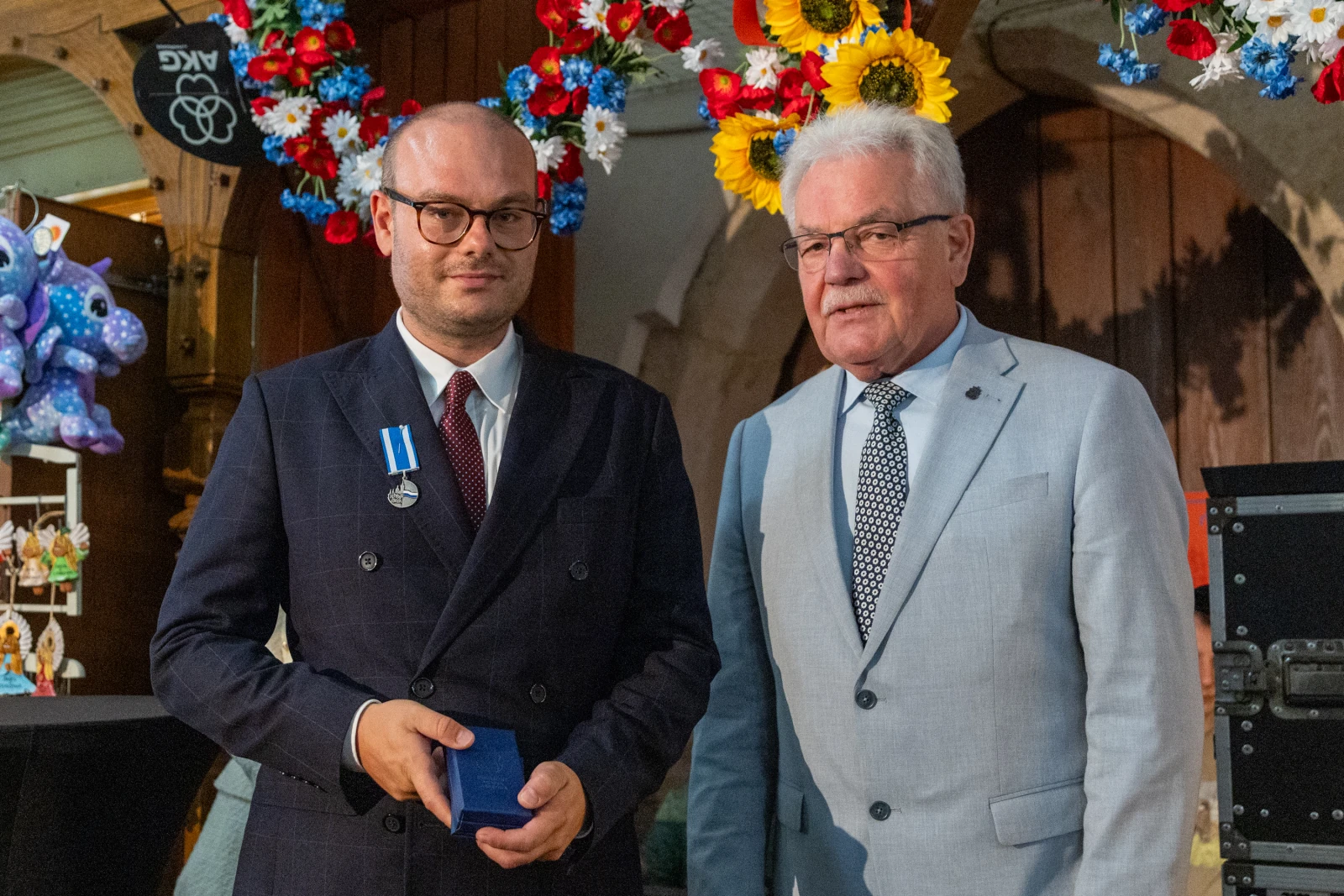Mężczyzna w garniturze odbierający medal od innego mężczyzny w jasnym garniturze. W tle dekoracje kwiatowe.  Autor: Bogusław Świerzowski