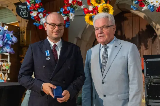 Mężczyzna w garniturze odbierający medal od innego mężczyzny w jasnym garniturze. W tle dekoracje kwiatowe.