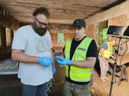 Dwóch mężczyzn w rękawiczkach trzyma fragment znaleziska archeologicznego w pomieszczeniu.