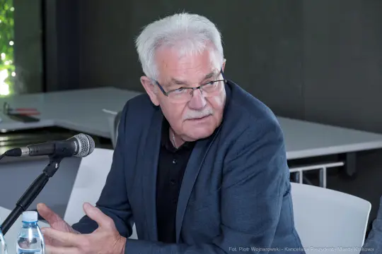 Mężczyzna z siwymi włosami siedzący przy stole, rozmawia do mikrofonu.