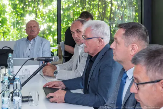 Mężczyźni w garniturach siedzą przy stole, prowadzą dyskusję w otoczeniu zieleni.