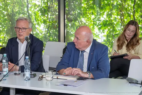 Dwaj mężczyźni rozmawiają przy stole, w tle kobieta pisząca notatki. Zielone rośliny w tle.