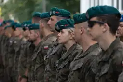 Żołnierze w mundurach wojskowych z beretami w kolejce, w tle zieleń.