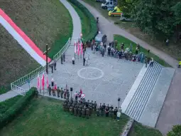 Uroczystość z flagami, żołnierzami i zgromadzonymi ludźmi na placu.